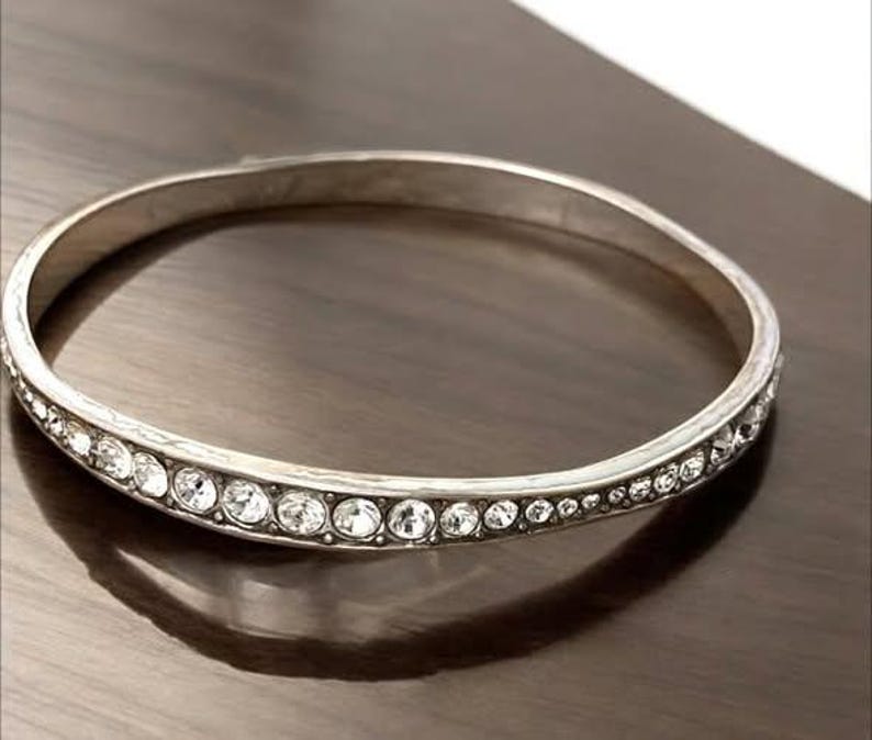 Puede incluir: Una pulsera r&iacute;gida de metal en tono plateado que presenta una hilera continua de cristales transparentes de corte redondo incrustados en la superficie exterior. La pulsera tiene un acabado liso y pulido con una forma circular.