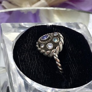 May include: A silver-toned ring with a cluster of small, colorful gemstones. The ring's setting has a textured, ornate design, and the band is twisted. The ring is displayed in a clear, square container with a black velvet interior.