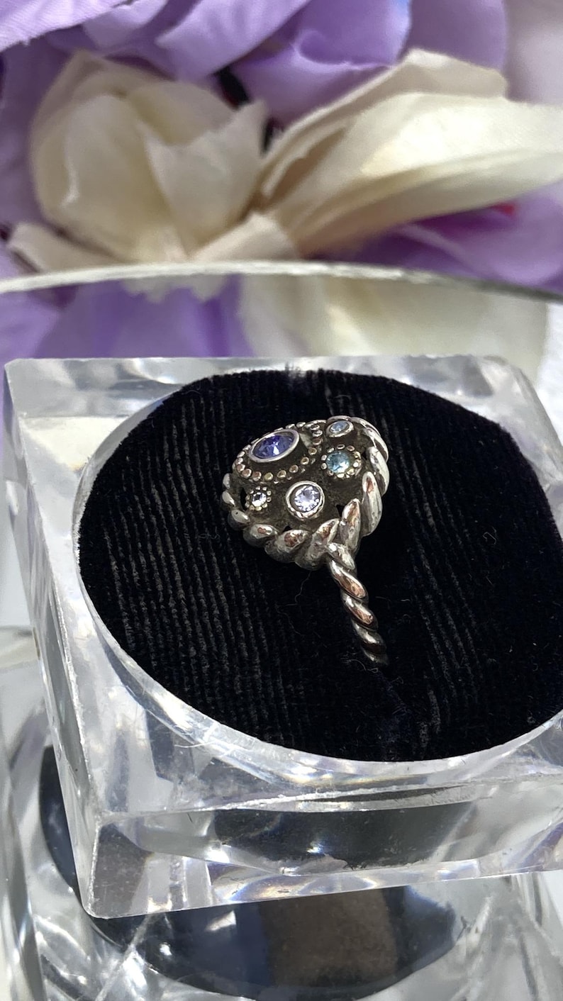 May include: A silver-toned ring with a cluster of small, colorful gemstones. The ring's setting has a textured, ornate design, and the band is twisted. The ring is displayed in a clear, square container with a black velvet interior.