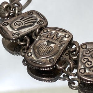 Puede incluir: Una pulsera de eslabones de metal plateado con dijes rectangulares de dise&ntilde;os en relieve. Los dijes muestran una mano con una espiral, un coraz&oacute;n coronado y un motivo floral de cuatro p&eacute;talos. Los eslabones est&aacute;n unidos por anillos circulares.