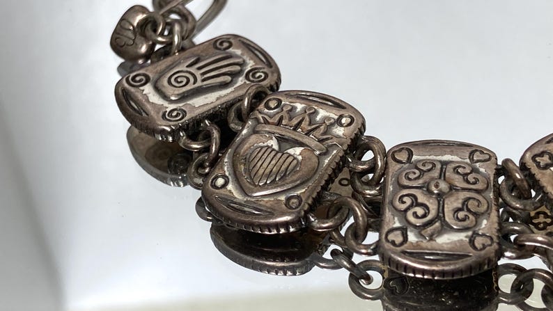 Puede incluir: Una pulsera de eslabones de metal plateado con dijes rectangulares de dise&ntilde;os en relieve. Los dijes muestran una mano con una espiral, un coraz&oacute;n coronado y un motivo floral de cuatro p&eacute;talos. Los eslabones est&aacute;n unidos por anillos circulares.
