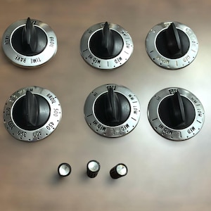 May include: A set of six silver and black oven control knobs. The knobs have black handles and silver faces with markings for temperature and cooking functions like "Bake" and "Broil". Three smaller silver and black knobs are also present.