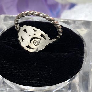 May include: A silver ring with a floral design and a twisted band. The ring is set on a black velvet surface inside a clear, square display case. The ring has a small, engraved emblem.