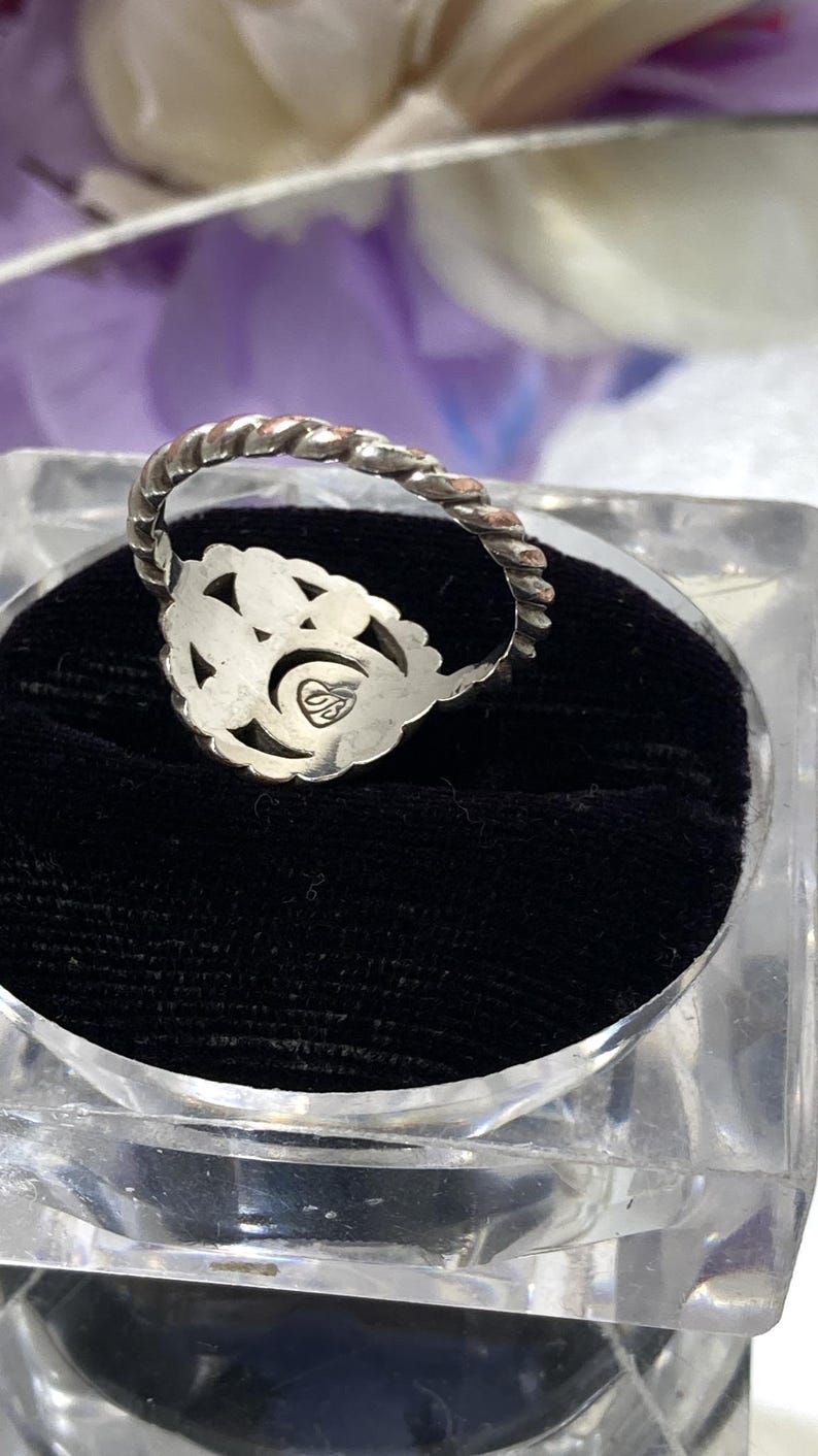 May include: A silver ring with a floral design and a twisted band. The ring is set on a black velvet surface inside a clear, square display case. The ring has a small, engraved emblem.