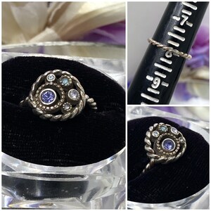 May include: A silver-toned ring with a celestial design. The ring features a central, round, dark blue gemstone surrounded by smaller, light blue and clear gemstones. The band has a twisted rope design.