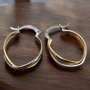 May include: A pair of oval hoop earrings with a two-tone design. The earrings feature a silver-colored outer band and a gold-colored inner band. The earrings are displayed on a dark brown wooden surface.