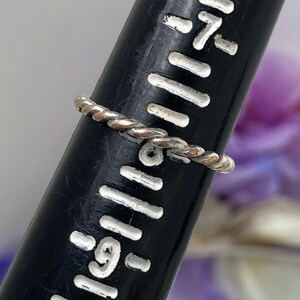 May include: A silver-colored, twisted rope ring is displayed on a black ring sizer. The ring is thin and delicate, with a braided design. The ring sizer has white markings for ring sizes.