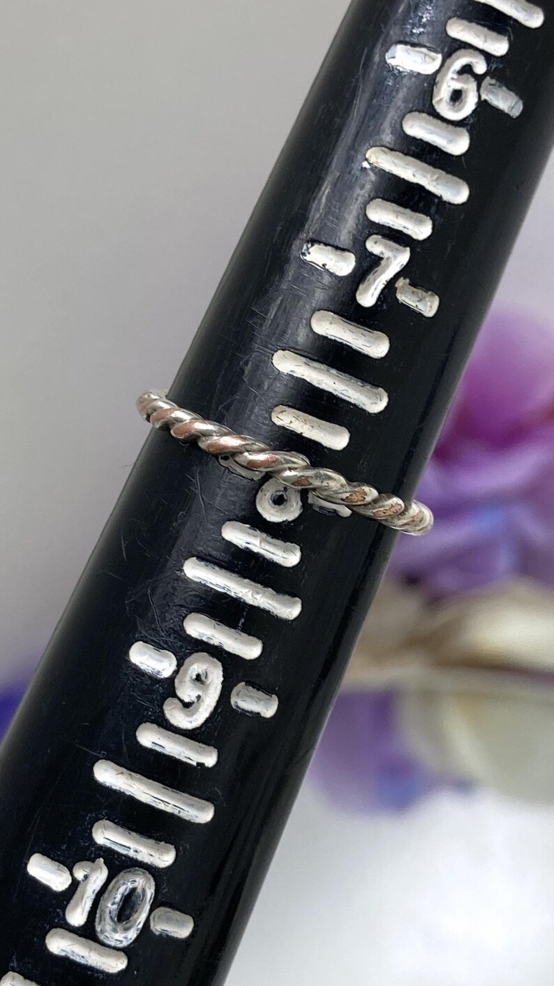 May include: A silver-colored, twisted rope ring is displayed on a black ring sizer. The ring is thin and delicate, with a braided design. The ring sizer has white markings for ring sizes.