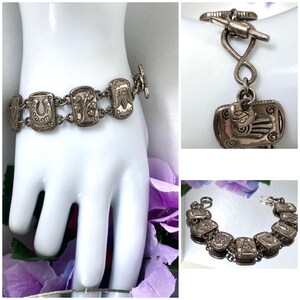 Puede incluir: Una pulsera de eslabones en tono plateado con dijes rectangulares que presentan dise&ntilde;os en relieve, incluyendo una herradura, un &aacute;rbol, un coraz&oacute;n y una mano. La pulsera se asegura con un cierre de barra y tiene un acabado met&aacute;lico desgastado.