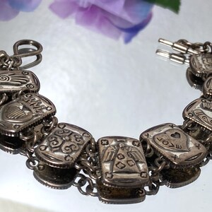 Puede incluir: Una pulsera de eslabones en tono plateado con una serie de dijes rectangulares, cada uno grabado con s&iacute;mbolos distintos que incluyen una mano, un coraz&oacute;n con la palabra LOVE, una herradura y varios patrones geom&eacute;tricos, unidos por anillas.