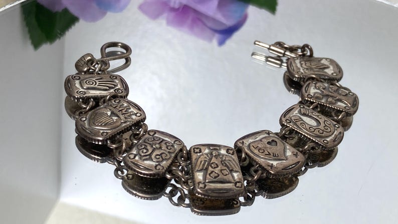 Puede incluir: Una pulsera de eslabones en tono plateado con una serie de dijes rectangulares, cada uno grabado con s&iacute;mbolos distintos que incluyen una mano, un coraz&oacute;n con la palabra LOVE, una herradura y varios patrones geom&eacute;tricos, unidos por anillas.