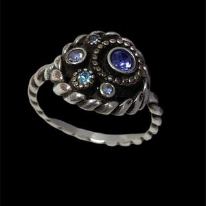 May include: A silver ring with a textured band and a cluster of blue gemstones. The ring's centerpiece features a large, round, blue stone surrounded by smaller blue and light blue stones, set in a dark, textured setting.