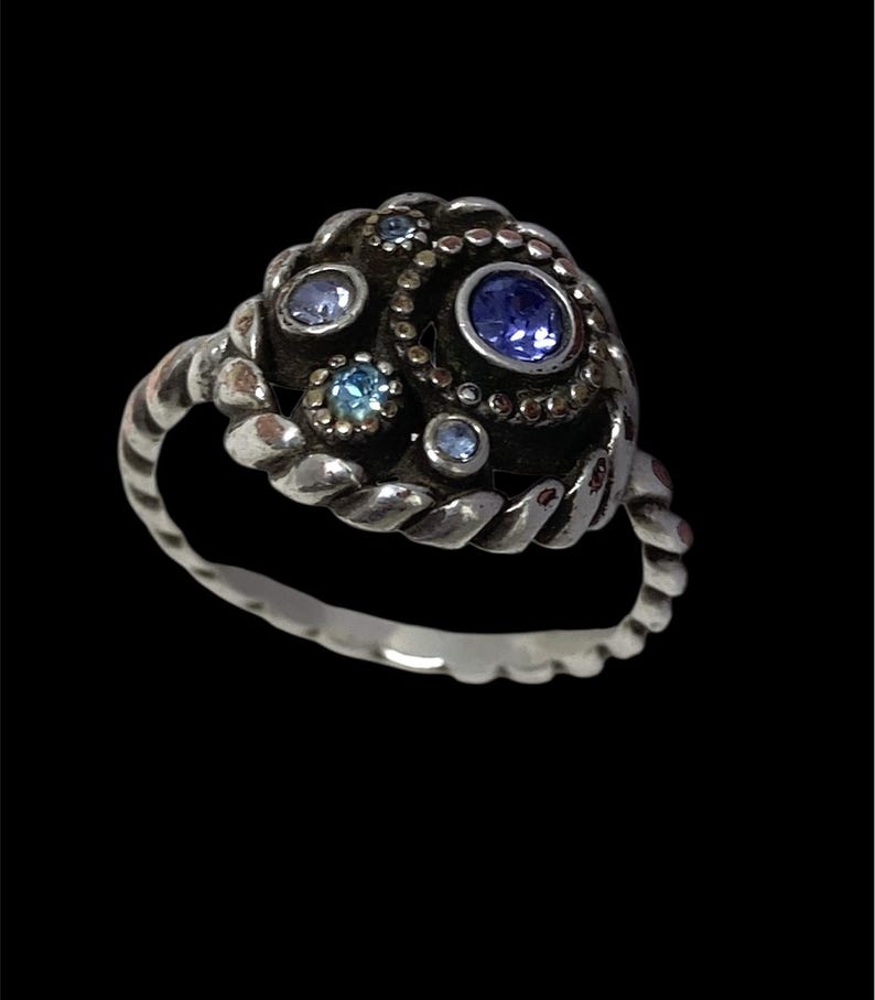May include: A silver ring with a textured band and a cluster of blue gemstones. The ring's centerpiece features a large, round, blue stone surrounded by smaller blue and light blue stones, set in a dark, textured setting.