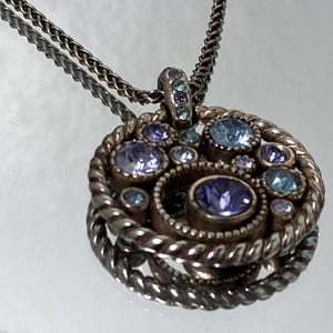 K&ouml;nnte beinhalten: Eine kreisf&ouml;rmige Anh&auml;ngerkette mit einem gedrehten Metallrahmen und einer Ansammlung runder, unterschiedlich gro&szlig;er Kristalle in Hellblau, Lavendel und Violett. Der Anh&auml;nger h&auml;ngt an einer d&uuml;nnen, strukturierten silberfarbenen Metallkette.
