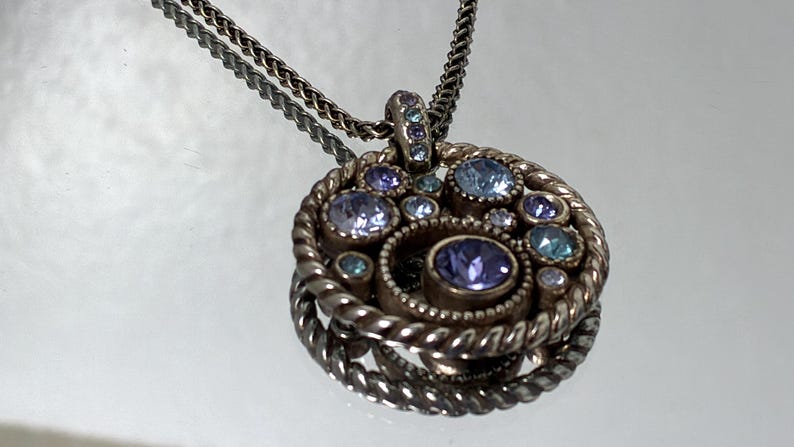 K&ouml;nnte beinhalten: Eine kreisf&ouml;rmige Anh&auml;ngerkette mit einem gedrehten Metallrahmen und einer Ansammlung runder, unterschiedlich gro&szlig;er Kristalle in Hellblau, Lavendel und Violett. Der Anh&auml;nger h&auml;ngt an einer d&uuml;nnen, strukturierten silberfarbenen Metallkette.