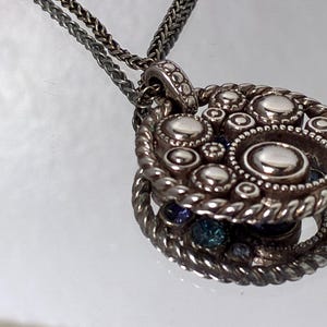 K&ouml;nnte beinhalten: Eine silberfarbene Anh&auml;ngerkette mit kreisf&ouml;rmigem Design, erhabenen, polierten Metallkugeln und einem gedrehten seilartigen Rand. Der Anh&auml;nger h&auml;ngt an einer feinen, geflochtenen silberfarbenen Kette und weist eine reflektierende Oberfl&auml;che auf.