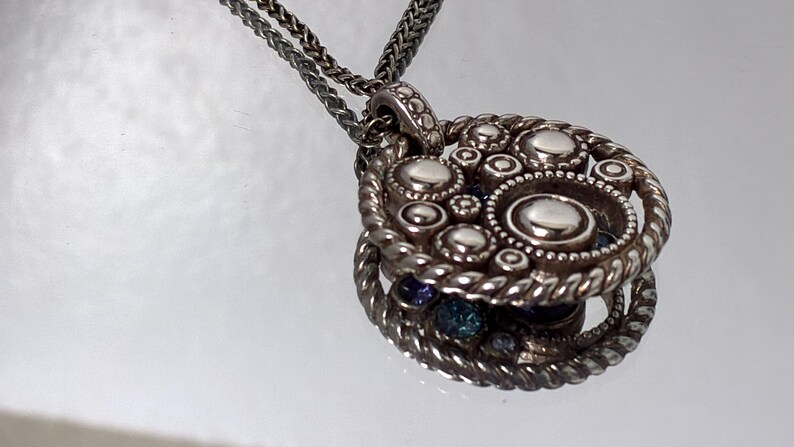 K&ouml;nnte beinhalten: Eine silberfarbene Anh&auml;ngerkette mit kreisf&ouml;rmigem Design, erhabenen, polierten Metallkugeln und einem gedrehten seilartigen Rand. Der Anh&auml;nger h&auml;ngt an einer feinen, geflochtenen silberfarbenen Kette und weist eine reflektierende Oberfl&auml;che auf.