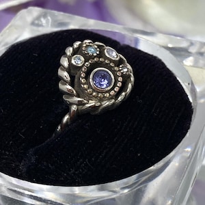 May include: A silver ring with a spiral design, featuring a central purple gemstone surrounded by smaller blue and clear stones. The ring is displayed on a black velvet surface inside a clear, square container.