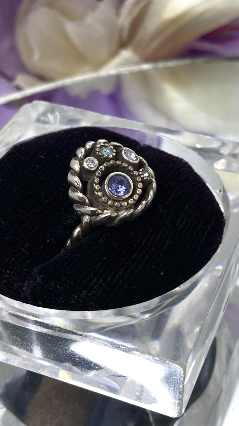 May include: A silver ring with a spiral design, featuring a central purple gemstone surrounded by smaller blue and clear stones. The ring is displayed on a black velvet surface inside a clear, square container.