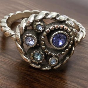 May include: A silver ring with a detailed design featuring a central cluster of blue and purple gemstones. The ring has a braided band and a decorative centerpiece with multiple smaller stones.