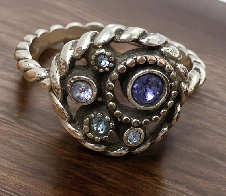 May include: A silver ring with a detailed design featuring a central cluster of blue and purple gemstones. The ring has a braided band and a decorative centerpiece with multiple smaller stones.