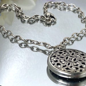 May include: Silver-toned necklace with a large, round pendant. The pendant features an intricate, openwork design over a black background. The chain is a chunky link style, and the necklace is displayed on a reflective surface.