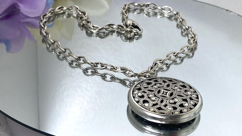 May include: Silver-toned necklace with a large, round pendant. The pendant features an intricate, openwork design over a black background. The chain is a chunky link style, and the necklace is displayed on a reflective surface.