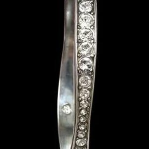 Puede incluir: Una pulsera r&iacute;gida de tono plateado que presenta una fila continua de cristales transparentes de corte redondo a lo largo del borde exterior. La banda met&aacute;lica tiene un acabado liso y pulido en el interior y un perfil ligeramente ondulado.