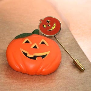 May include: A bright orange pumpkin brooch with a green leaf and a carved face featuring triangular eyes and a wide grin, accompanied by a thin gold-toned metal stick pin topped with a small, circular red pumpkin charm with a smiling face.