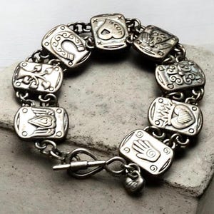 Puede incluir: Una pulsera de dijes en tono plateado con una serie de eslabones rectangulares, cada uno grabado con s&iacute;mbolos distintos que incluyen una herradura, un coraz&oacute;n, una mano y alas. La pulsera se asegura con un cierre de barra y un peque&ntilde;o dije de coraz&oacute;n.