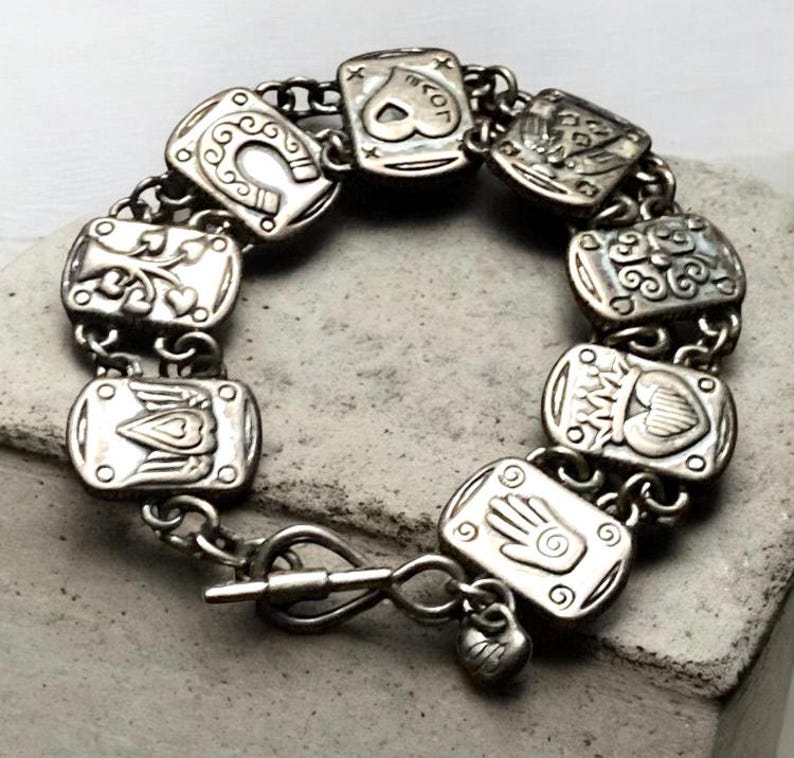 Puede incluir: Una pulsera de dijes en tono plateado con una serie de eslabones rectangulares, cada uno grabado con s&iacute;mbolos distintos que incluyen una herradura, un coraz&oacute;n, una mano y alas. La pulsera se asegura con un cierre de barra y un peque&ntilde;o dije de coraz&oacute;n.