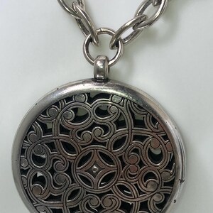 May include: Silver-toned necklace featuring a large, round pendant with an intricate openwork design. The pendant hangs from a chunky chain. The detailed pattern within the pendant is visible against a dark background.