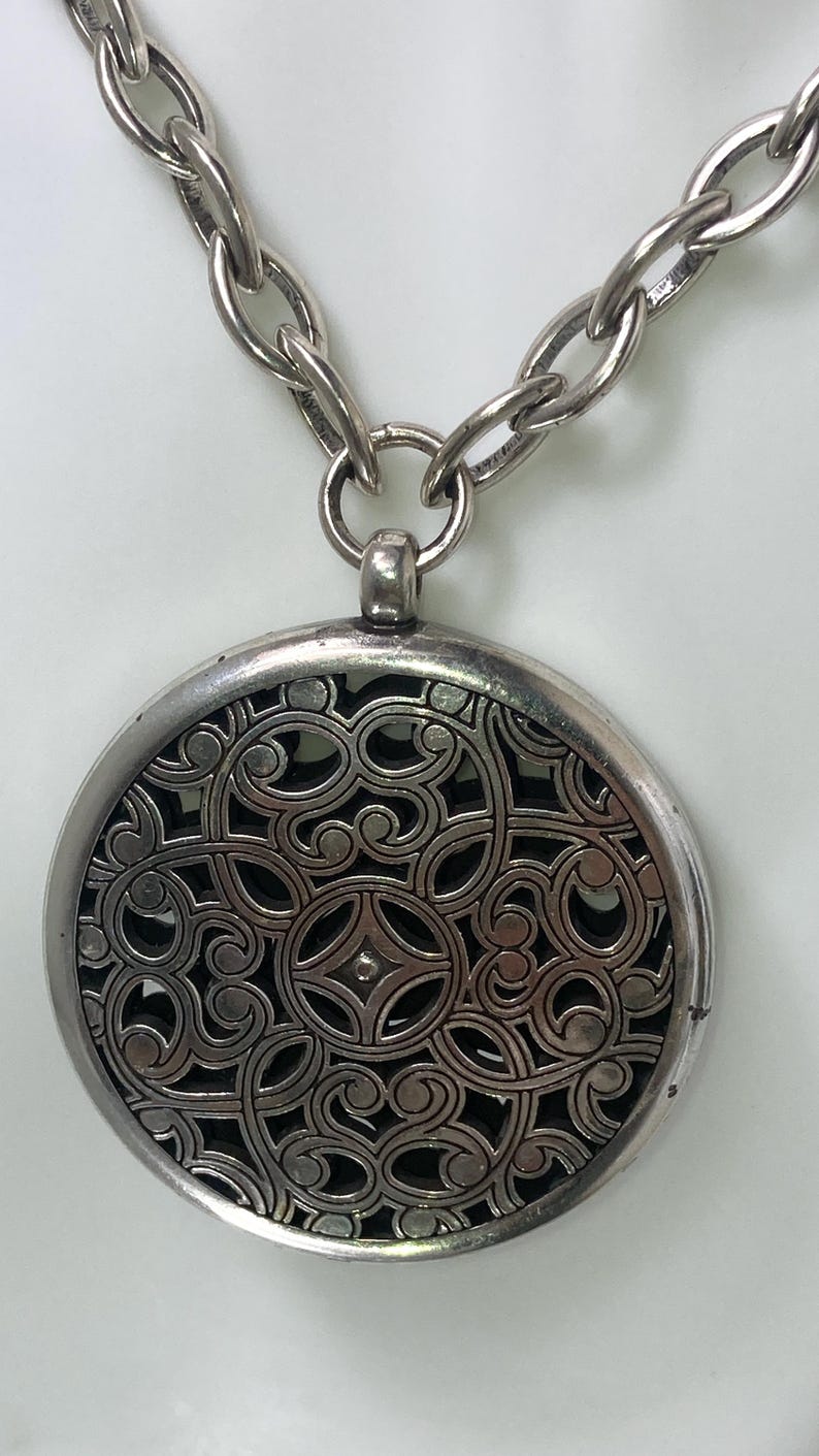May include: Silver-toned necklace featuring a large, round pendant with an intricate openwork design. The pendant hangs from a chunky chain. The detailed pattern within the pendant is visible against a dark background.