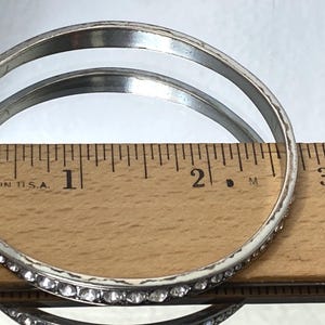 Puede incluir: Un brazalete de metal en tono plateado con una hilera de peque&ntilde;os diamantes de imitaci&oacute;n transparentes de corte redondo. El brazalete est&aacute; colocado contra una regla de madera, mostrando un di&aacute;metro interior de unos 6,4 cm.