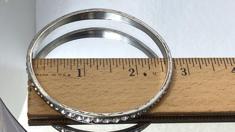 Puede incluir: Un brazalete de metal en tono plateado con una hilera de peque&ntilde;os diamantes de imitaci&oacute;n transparentes de corte redondo. El brazalete est&aacute; colocado contra una regla de madera, mostrando un di&aacute;metro interior de unos 6,4 cm.