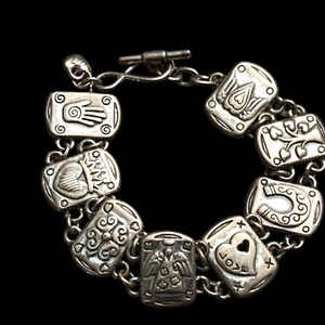 Puede incluir: Una pulsera de dijes en tono plateado con una serie de eslabones rectangulares, cada uno grabado con s&iacute;mbolos distintos que incluyen una mano, un coraz&oacute;n, un &aacute;rbol, una herradura y un &aacute;ngel. La pulsera se asegura con un cierre de barra.