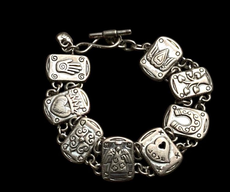 Puede incluir: Una pulsera de dijes en tono plateado con una serie de eslabones rectangulares, cada uno grabado con s&iacute;mbolos distintos que incluyen una mano, un coraz&oacute;n, un &aacute;rbol, una herradura y un &aacute;ngel. La pulsera se asegura con un cierre de barra.