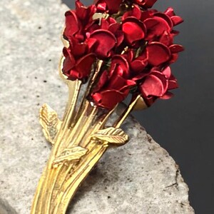 May include: A gold-tone metal brooch shaped like a bouquet of deep red roses. The stems are textured and gold-colored, featuring small gold leaves attached to the base. The rose petals are painted in a vibrant, glossy red finish with layered details.