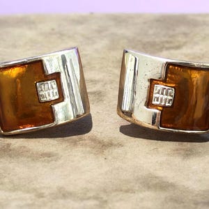 May include: A pair of rectangular earrings featuring a polished silver-tone metal frame with a curved surface. Each earring contains a smooth, translucent amber-colored resin inlay and a small, square silver-tone accent piece in the center.