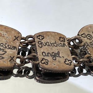 Puede incluir: Una pulsera de eslabones de metal plateado con dijes rectangulares grabados con texto y s&iacute;mbolos. Los dijes muestran las palabras Compassion, guardian angel y HARMONY, acompa&ntilde;adas de dise&ntilde;os grabados que incluyen una flor, corazones y espirales.