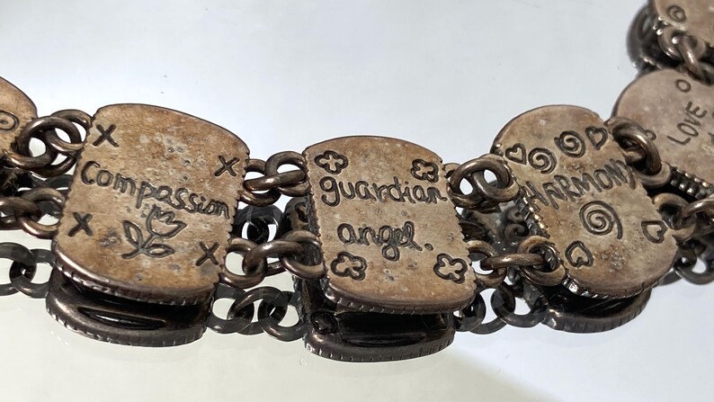 Puede incluir: Una pulsera de eslabones de metal plateado con dijes rectangulares grabados con texto y s&iacute;mbolos. Los dijes muestran las palabras Compassion, guardian angel y HARMONY, acompa&ntilde;adas de dise&ntilde;os grabados que incluyen una flor, corazones y espirales.