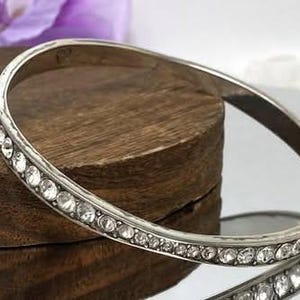 Puede incluir: Una pulsera r&iacute;gida de metal plateado que presenta una hilera continua de peque&ntilde;os diamantes de imitaci&oacute;n transparentes de corte redondo incrustados a lo largo de la superficie exterior. La pulsera tiene un acabado liso y pulido.