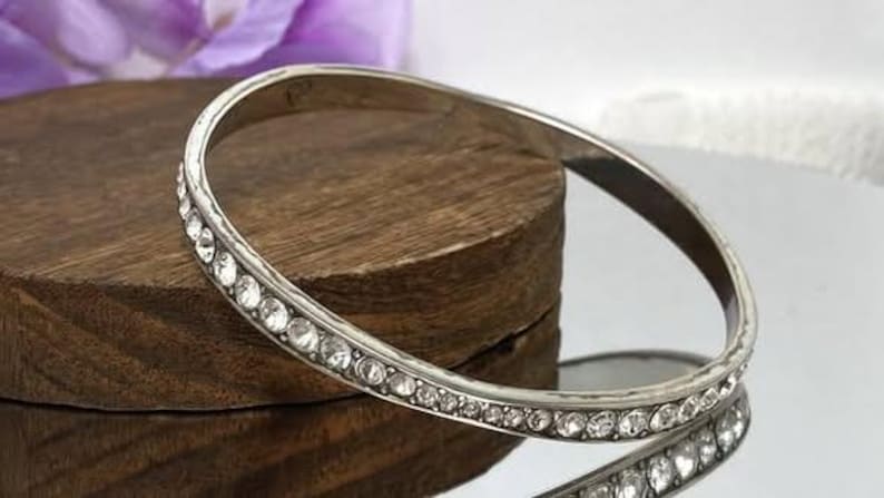 Puede incluir: Una pulsera r&iacute;gida de metal plateado que presenta una hilera continua de peque&ntilde;os diamantes de imitaci&oacute;n transparentes de corte redondo incrustados a lo largo de la superficie exterior. La pulsera tiene un acabado liso y pulido.