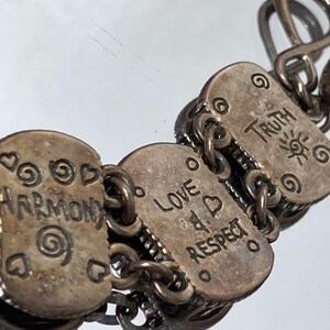 Puede incluir: Una pulsera de eslabones de metal plateado con dijes rectangulares grabados con las palabras Guardian, Harmony, Love, Respect y Truth, acompa&ntilde;ados de peque&ntilde;os motivos de espirales y corazones, unidos por anillas met&aacute;licas y un cierre de gancho.