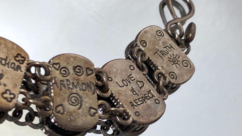 Puede incluir: Una pulsera de eslabones de metal plateado con dijes rectangulares grabados con las palabras Guardian, Harmony, Love, Respect y Truth, acompa&ntilde;ados de peque&ntilde;os motivos de espirales y corazones, unidos por anillas met&aacute;licas y un cierre de gancho.