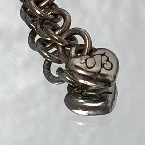 K&ouml;nnte beinhalten: Ein silberfarbenes Kettenarmband mit einer Reihe ineinandergreifender runder Glieder. Am Ende der Kette befinden sich zwei kleine, herzf&ouml;rmige Metallanh&auml;nger, wobei die Buchstaben OB deutlich auf der Oberfl&auml;che des oberen Herzens eingraviert sind.