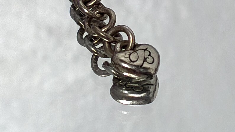 K&ouml;nnte beinhalten: Ein silberfarbenes Kettenarmband mit einer Reihe ineinandergreifender runder Glieder. Am Ende der Kette befinden sich zwei kleine, herzf&ouml;rmige Metallanh&auml;nger, wobei die Buchstaben OB deutlich auf der Oberfl&auml;che des oberen Herzens eingraviert sind.