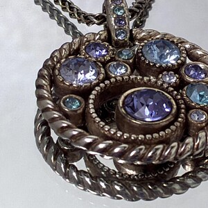 K&ouml;nnte beinhalten: Ein runder Anh&auml;nger mit einem gedrehten Metallrahmen und einer zentralen Ansammlung von runden, facettierten Kristallen in Hellblau, Violett und Lila. Der Anh&auml;nger h&auml;ngt an einer passenden Kette aus gedrehtem Metall.