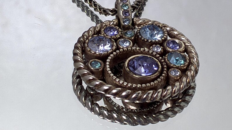K&ouml;nnte beinhalten: Ein runder Anh&auml;nger mit einem gedrehten Metallrahmen und einer zentralen Ansammlung von runden, facettierten Kristallen in Hellblau, Violett und Lila. Der Anh&auml;nger h&auml;ngt an einer passenden Kette aus gedrehtem Metall.