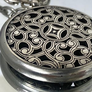 May include: A silver-toned, round pendant with an intricate, swirling, openwork design. The pendant has a black background and is attached to a silver chain. The design is symmetrical and detailed.